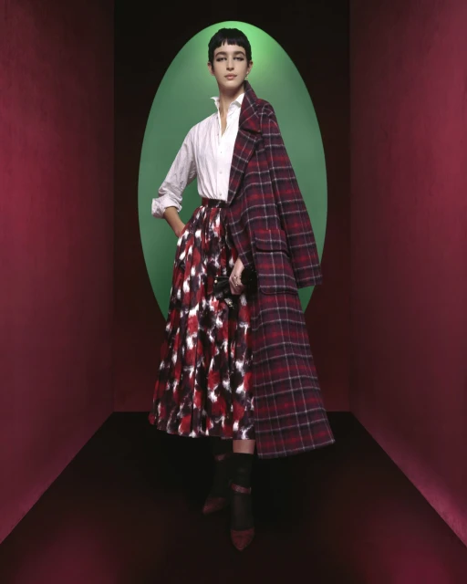 A woman in a white shirt and patterned skirt poses in front of a green oval, wearing a red plaid coat in a studio setting.
