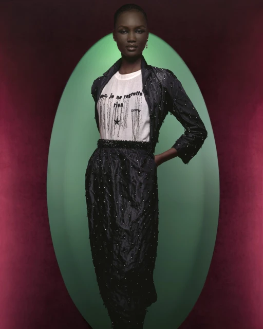 Dark-skinned model in dark skirt and jacket poses in front of an oval light-green background against dark red studio backdrop. Fashion shot.