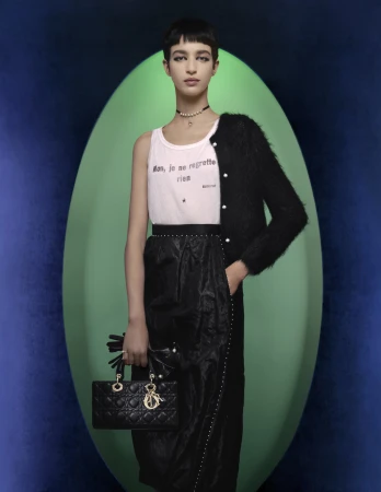 Fashion model holding a quilted black handbag, wearing a white slogan tank top, black cardigan, and skirt against an oval backdrop.
