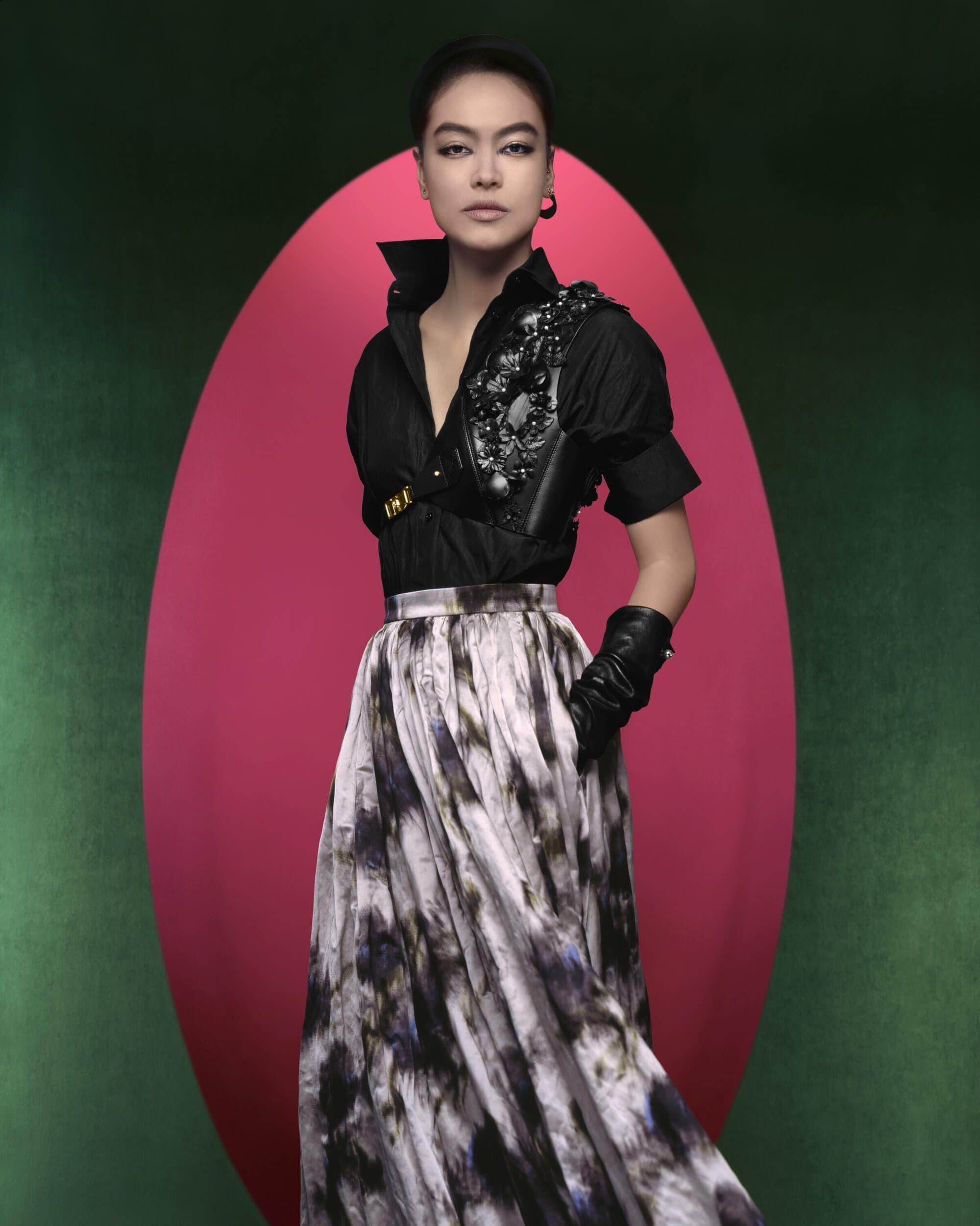 Portrait of a fashion model wearing a black button-up shirt, embellished harness, and full skirt against a pink and green backdrop.