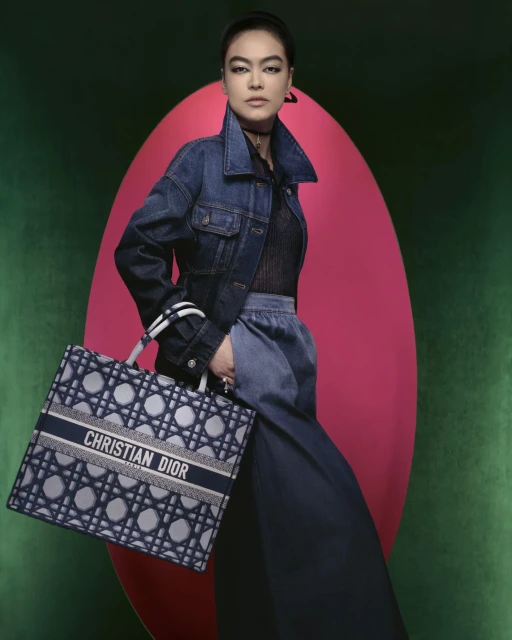 Fashion portrait of a woman wearing a denim jacket and holding a Christian Dior tote bag against a colorful studio backdrop.