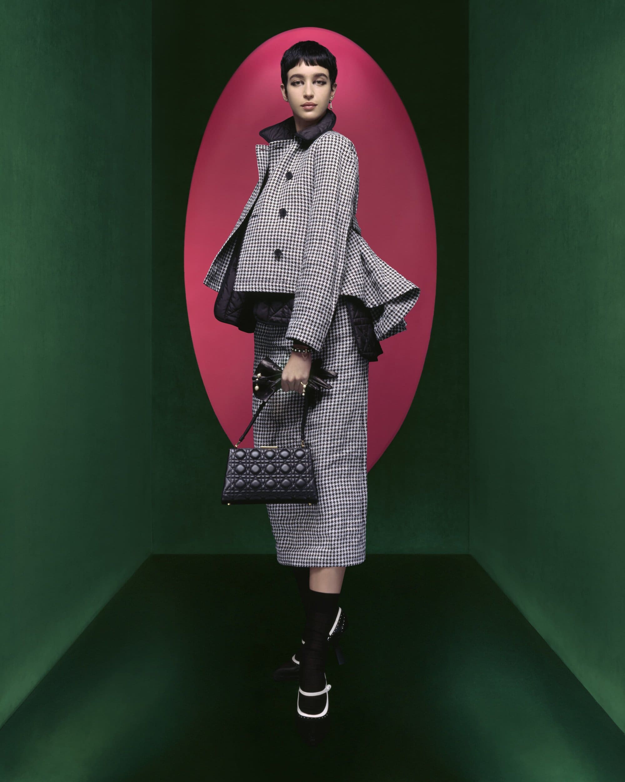 A fair-skinned young woman poses indoors wearing a black and white houndstooth skirt suit with a handbag, against a colorful background.