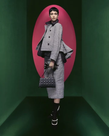 A fair-skinned young woman poses indoors wearing a black and white houndstooth skirt suit with a handbag, against a colorful background.