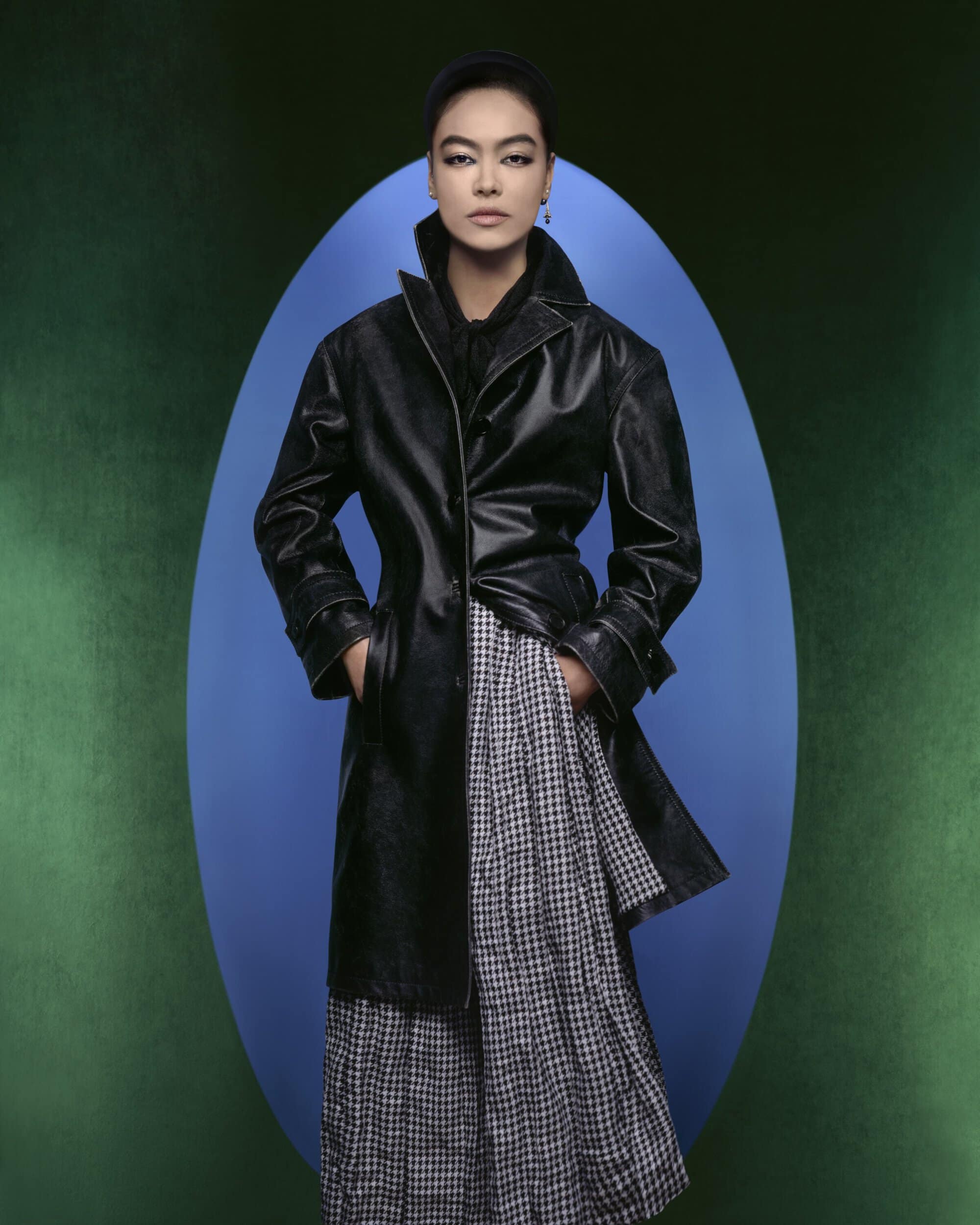 A woman in a black coat and checkered skirt poses in front of a blue oval against a green backdrop, in a fashion editorial shot.
