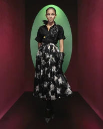 Model wearing a black blouse and patterned skirt standing in a set-dressed red and green arched room for Dior Fall 2023 campaign.