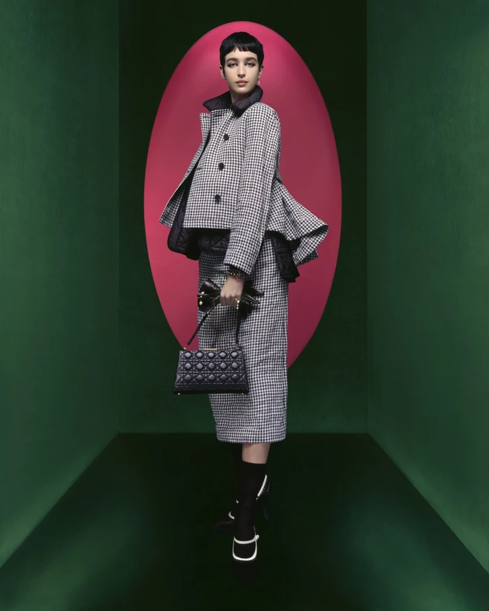 Model in houndstooth jacket and matching skirt holding a black handbag inside a green room with a pink oval backdrop