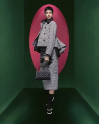 Model in houndstooth jacket and matching skirt holding a black handbag inside a green room with a pink oval backdrop