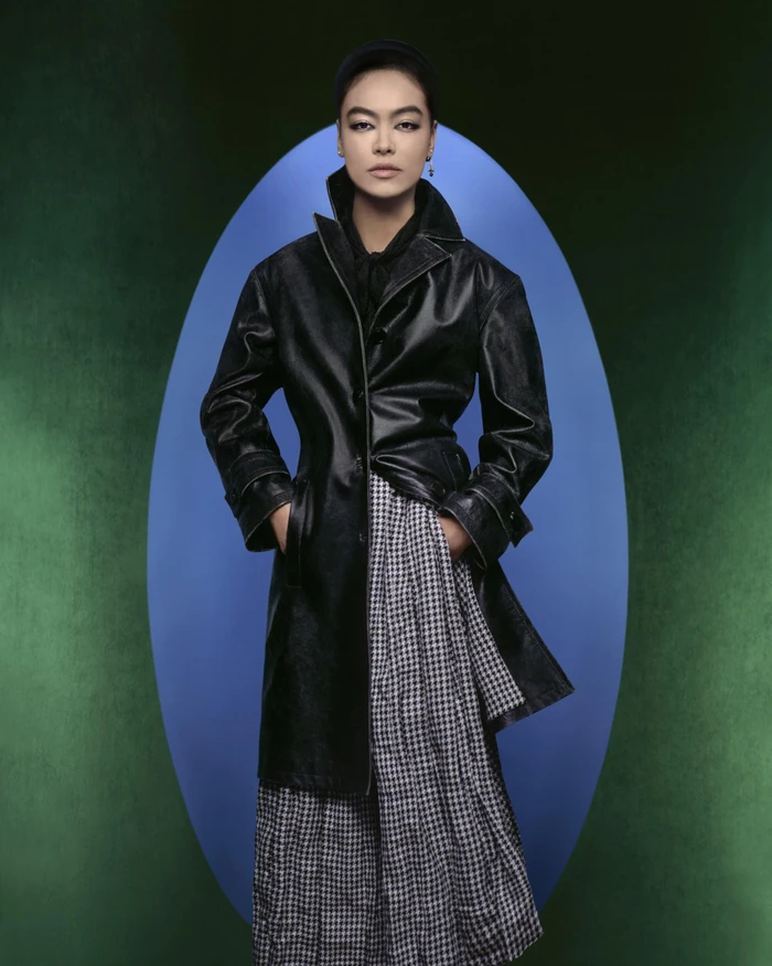 Model in a black leather coat and houndstooth skirt standing before an oval blue backdrop in a Dior fall campaign image.
