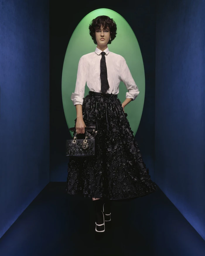 Model standing in front of an oval green archway wearing a white shirt, black tie, full black skirt, and holding a handbag.
