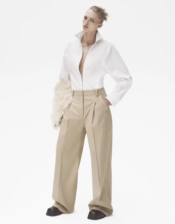 Fashion model in white shirt and tan wide-leg trousers poses in a studio with neutral tones, creating a minimalist and editorial aesthetic.