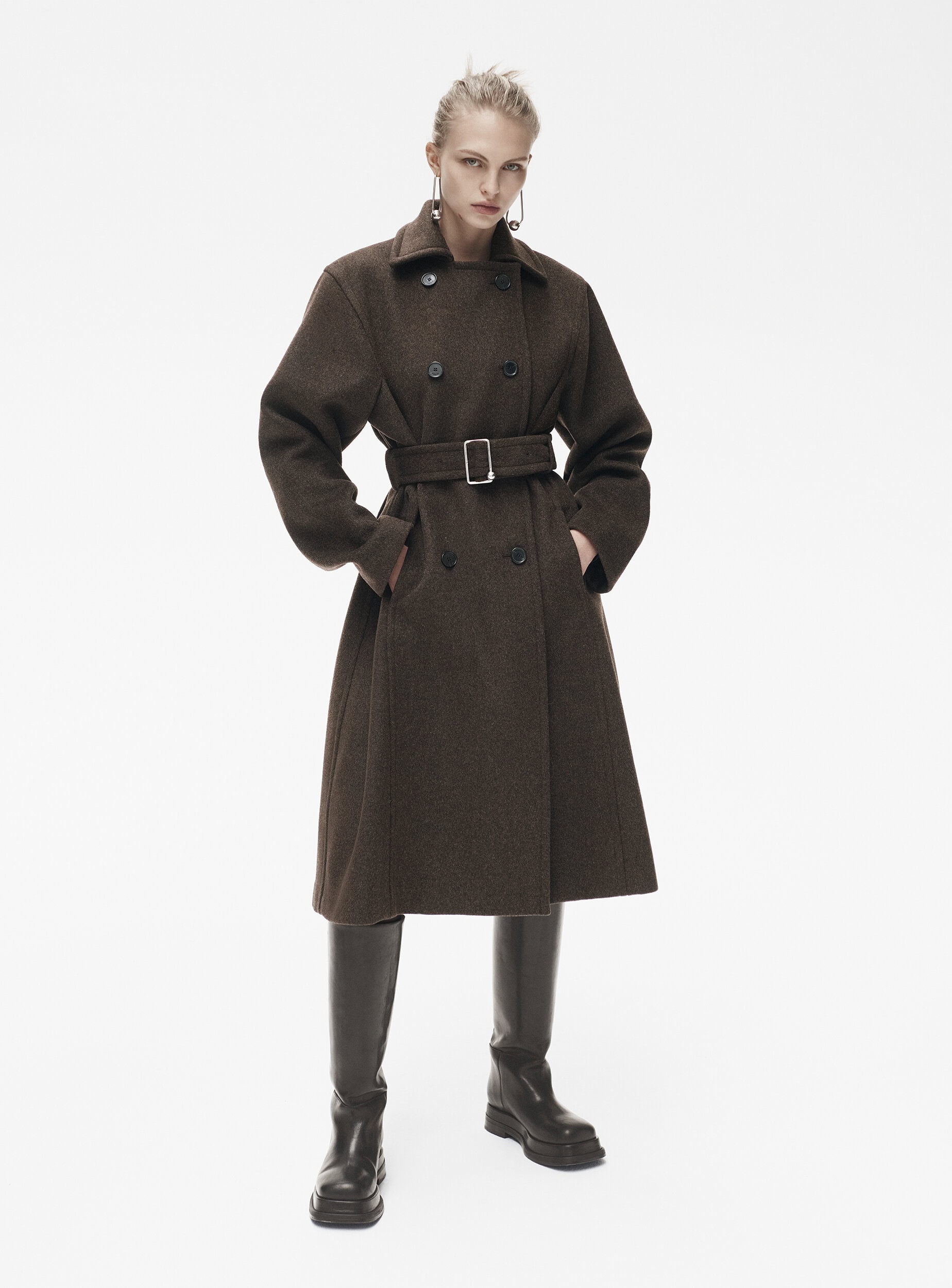 Full shot of a woman posing in a belted brown wool coat and knee-high leather boots in a bright photo studio setting with white background.