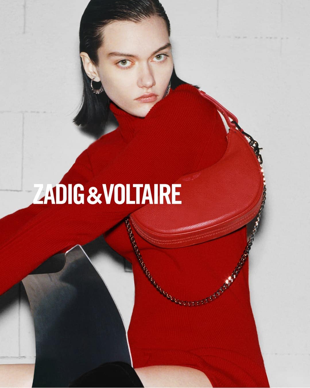 Fashion shot of a woman with short black hair wearing a red turtleneck, posing with a red purse, against a grey brick wall.