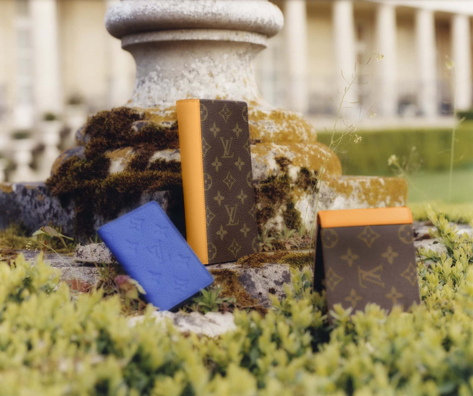 Three Louis Vuitton wallets are on a stone pedestal surrounded by green foliage, bathed in soft outdoor light with building in background.