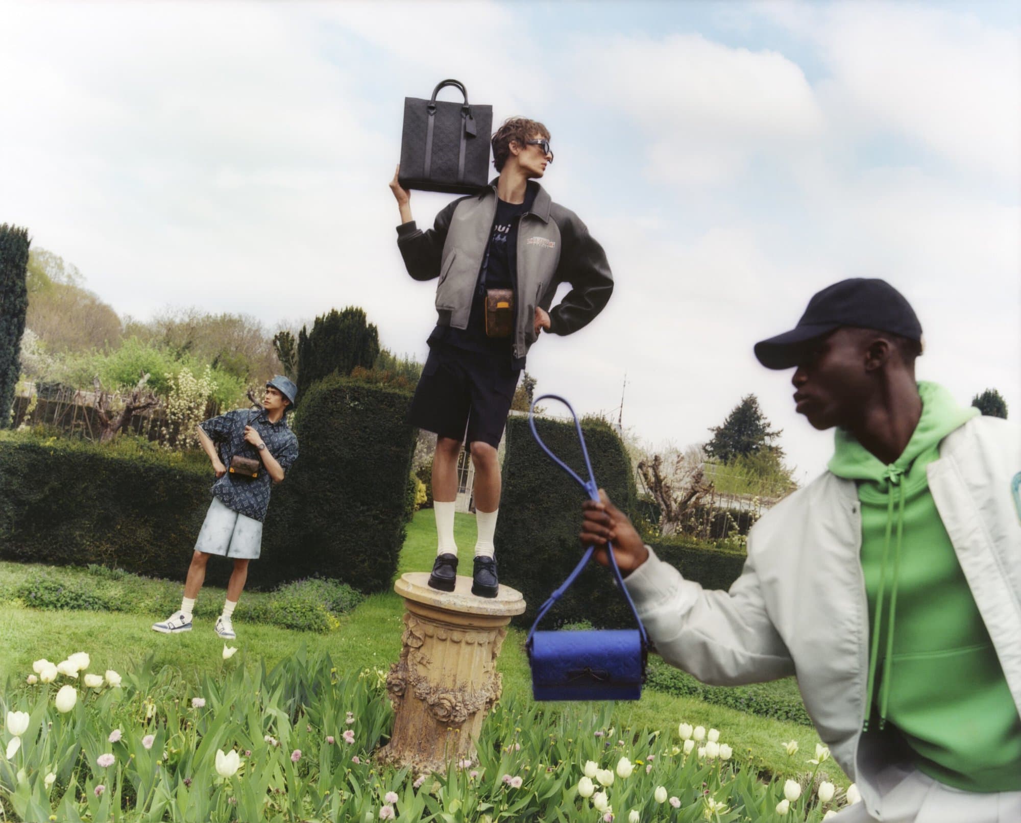 Three adult male fashion models pose with handbags in a garden with greenery and white flowers, showcasing menswear apparel and accessories.