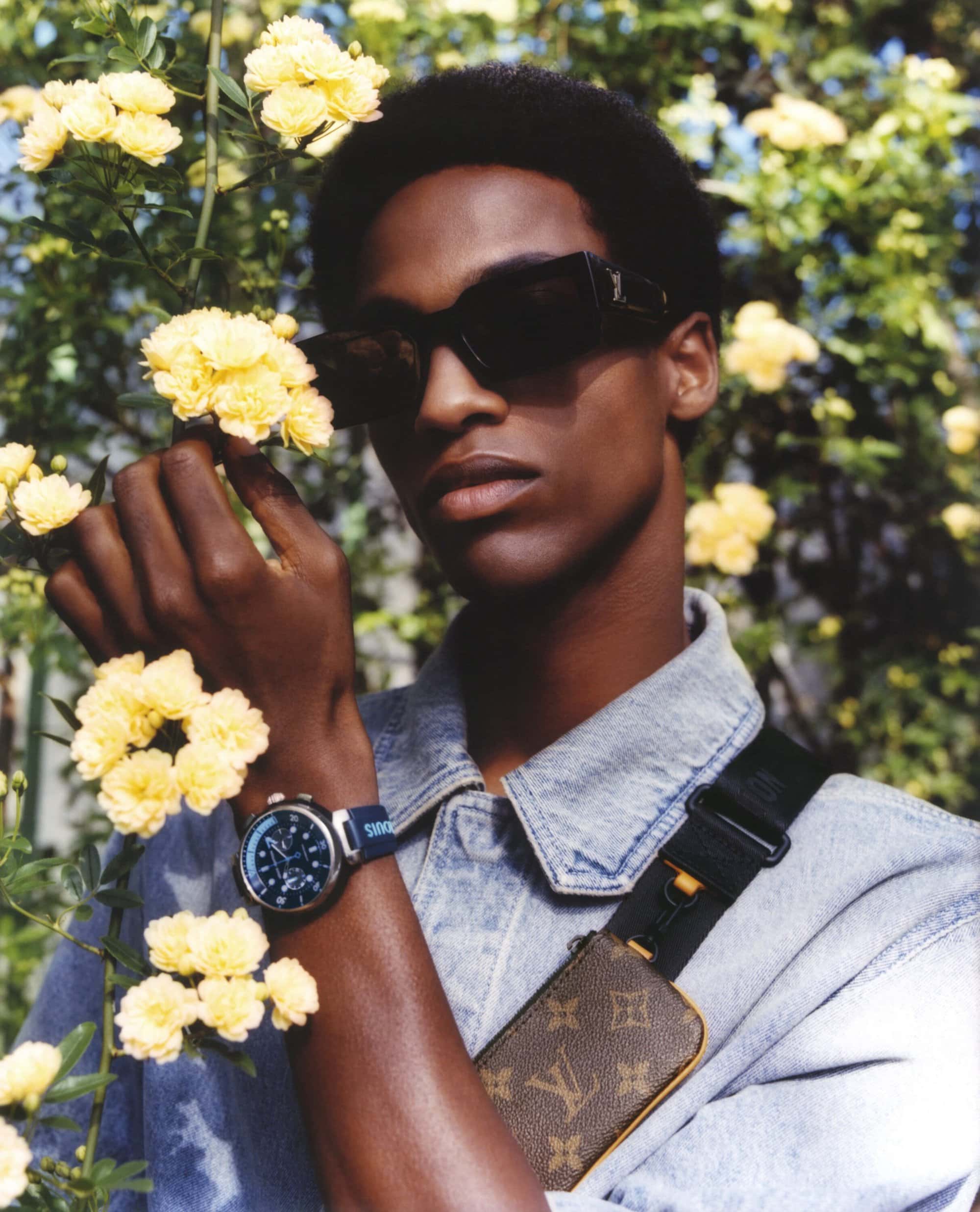 A stylish African-American man with sunglasses poses among yellow flowers, showcasing a designer shoulder bag and wristwatch.