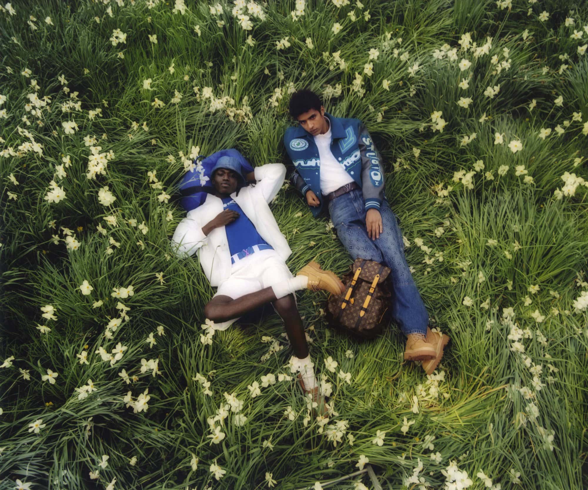 Two people lounge in a lush green field of small white flowers, wearing fashionable casual clothes, next to a brown monogrammed backpack.
