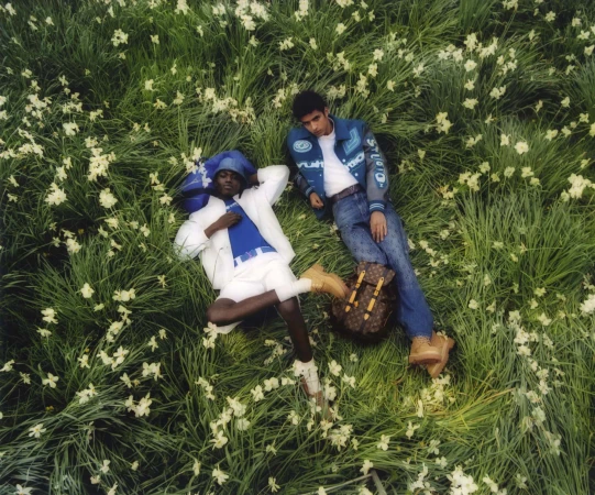 Two people lounge in a lush green field of small white flowers, wearing fashionable casual clothes, next to a brown monogrammed backpack.
