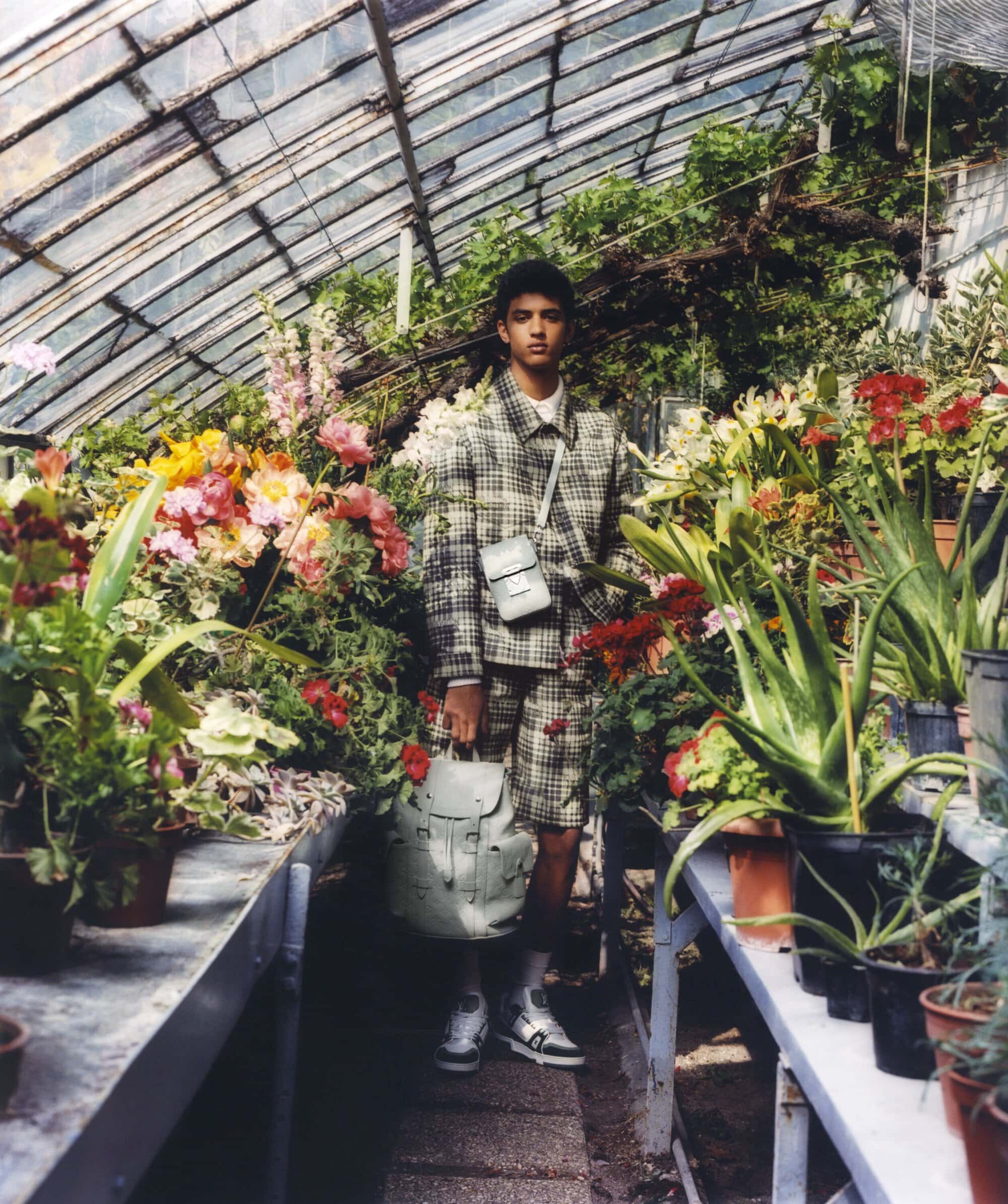 Man in a plaid suit standing inside a greenhouse filled with colorful flowers and potted plants, holding a backpack.