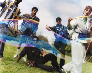 Male models wearing designer menswear outdoors on a green lawn posing around a large drifting iridescent soap bubble.