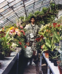 Model wearing a plaid jacket and shorts holding light green leather bags inside a lush glass greenhouse.