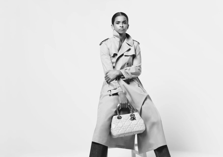 Black and white fashion portrait of an adult female in a trench coat, seated and holding a handbag, in a bright photo studio.