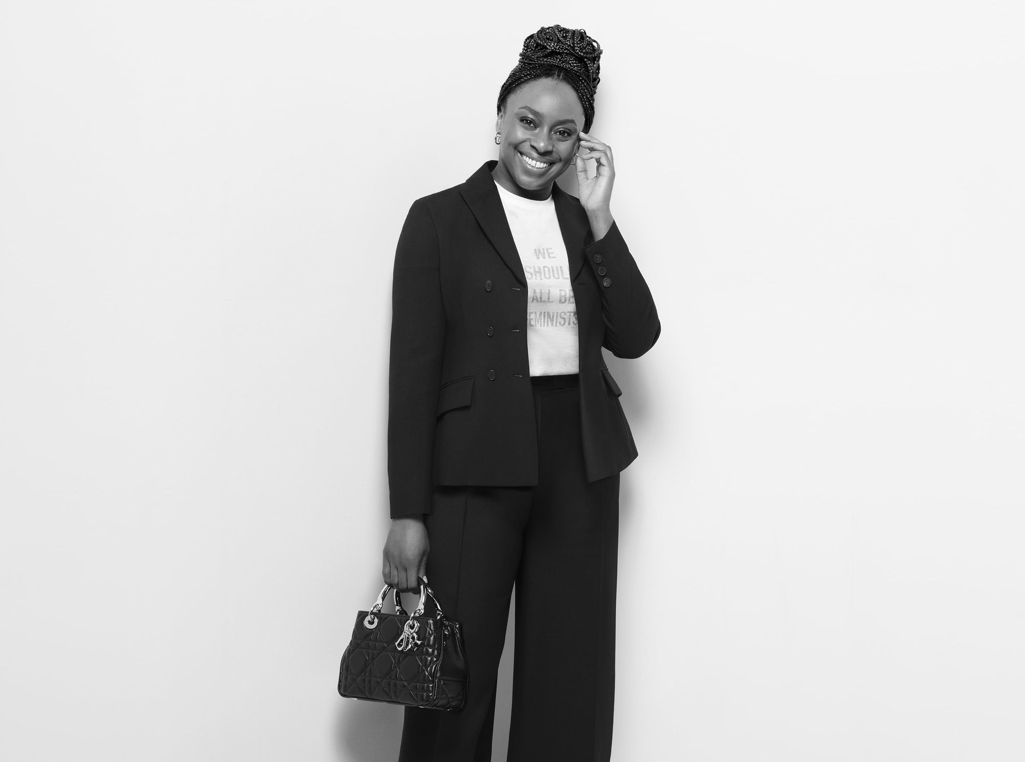 Black-and-white studio shot of a smiling adult female in a blazer and wide-leg pants, holding a Lady Dior handbag against a neutral backdrop.
