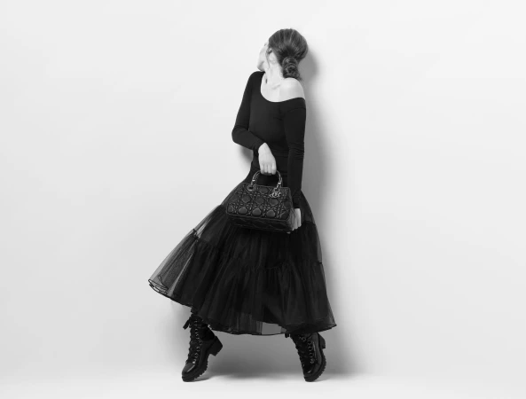 Black and white studio fashion shot of a woman in a black dress and boots, holding a Dior handbag, leaning against a white wall.