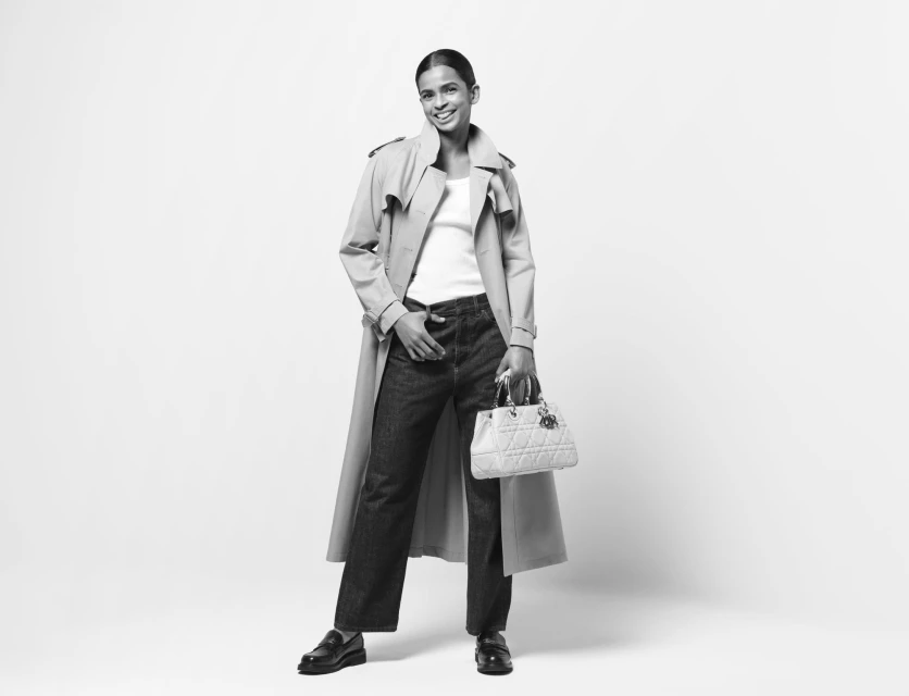 A full shot presents a smiling woman wearing a trench coat, jeans, and loafers, holding a white quilted handbag against a plain background.