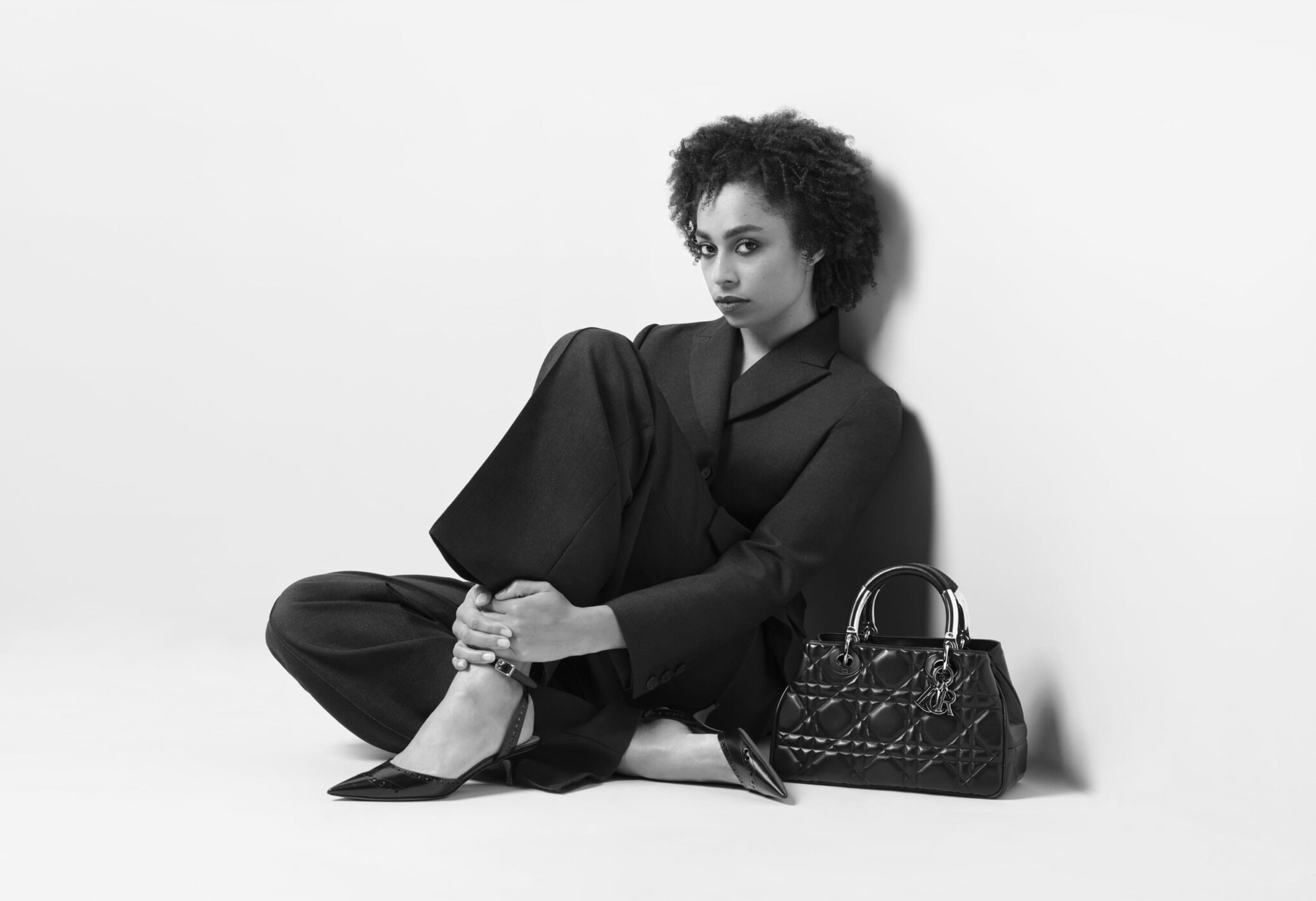 Black and white portrait of a young woman in a dark pantsuit sitting on the floor with a purse, shot in a photo studio setting.