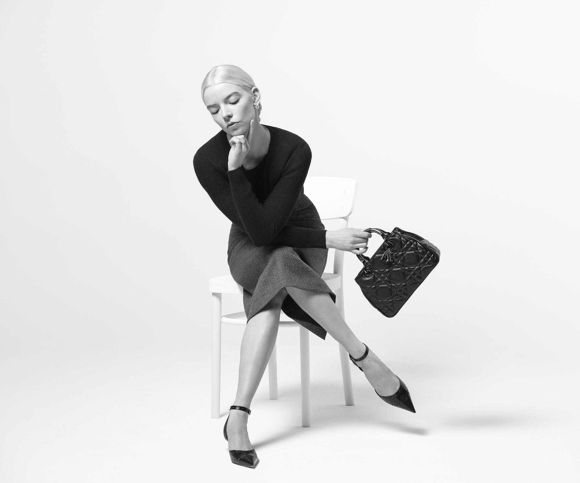 Anya Taylor-Joy seated on a white chair, wearing a black long-sleeved top and skirt, holding a quilted black handbag.