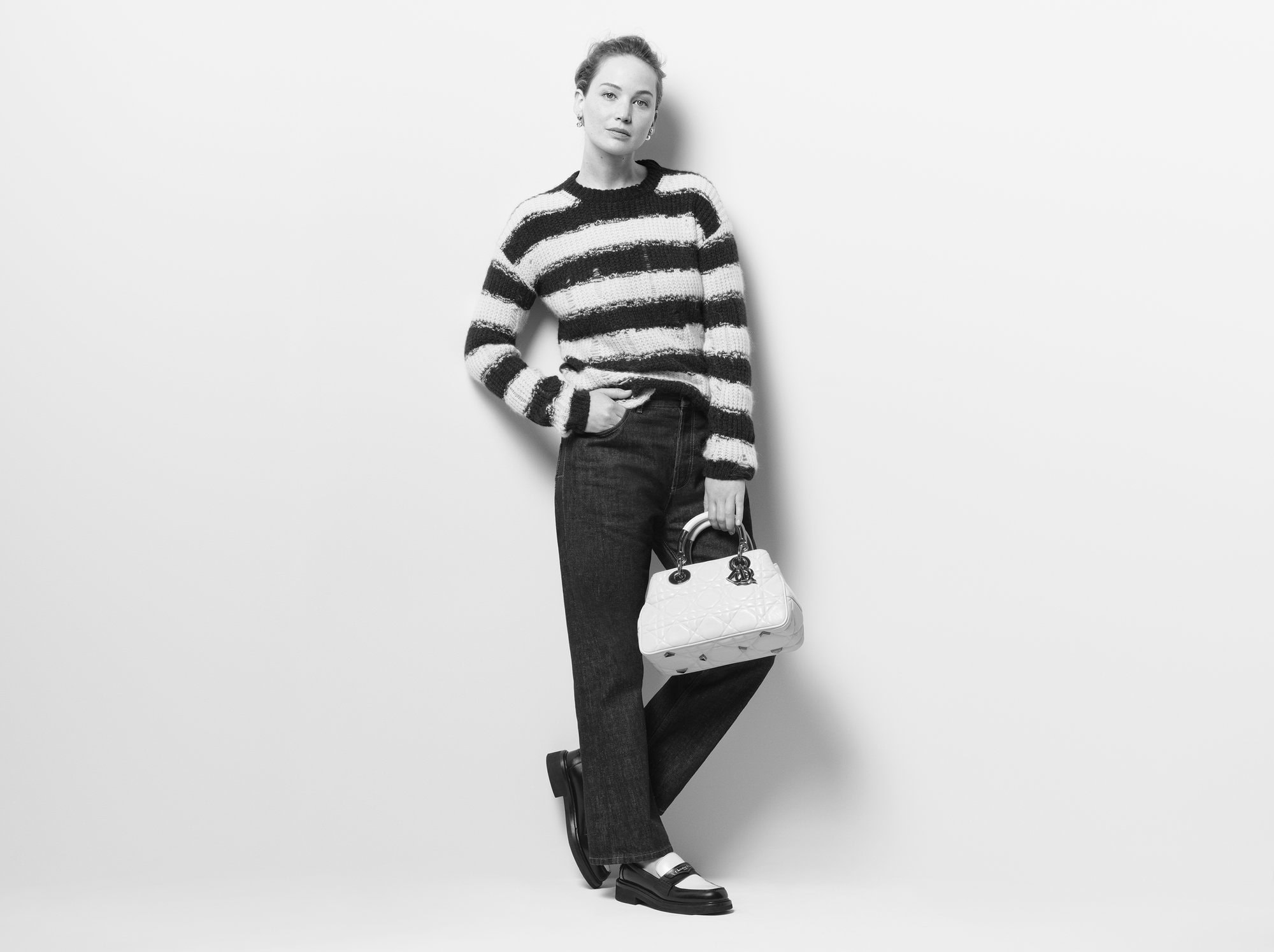 Monochrome full shot of a woman in a striped sweater and jeans holding a Dior purse, posing against a neutral background in a studio.