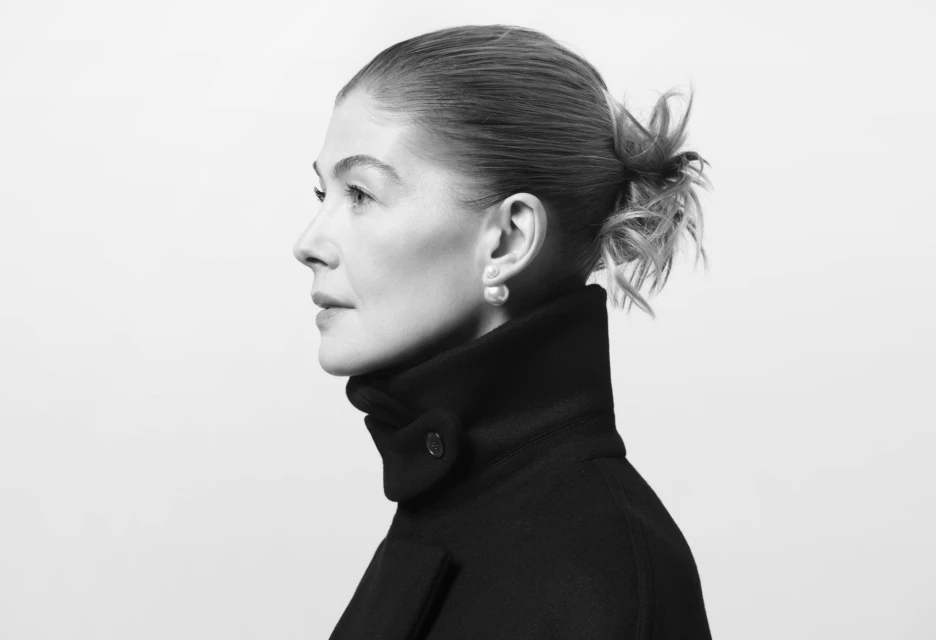 Black and white portrait of a woman in a dark coat, side profile, wearing pearl earrings against a light backdrop.
