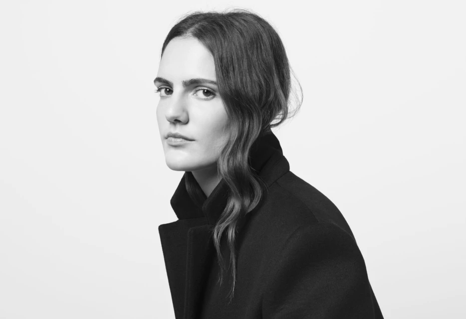 Monochrome portrait of an adult female model in a black overcoat looking over her shoulder against a bright white background.