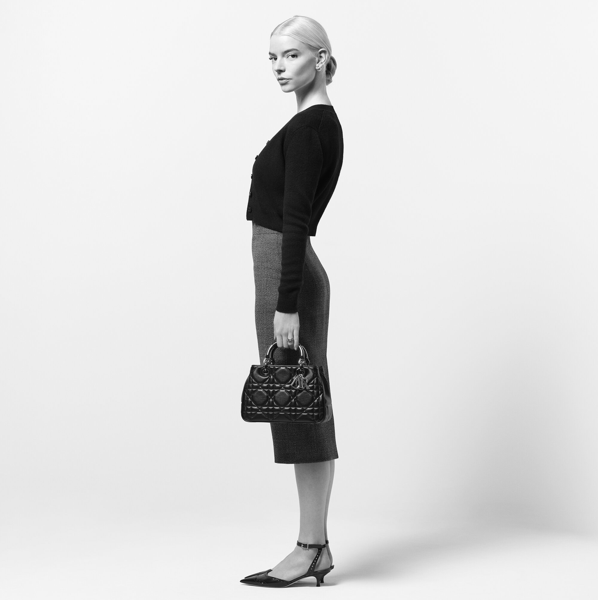 Anya Taylor-Joy in profile wearing a black top and midi skirt holding a black quilted Dior handbag against a white background.