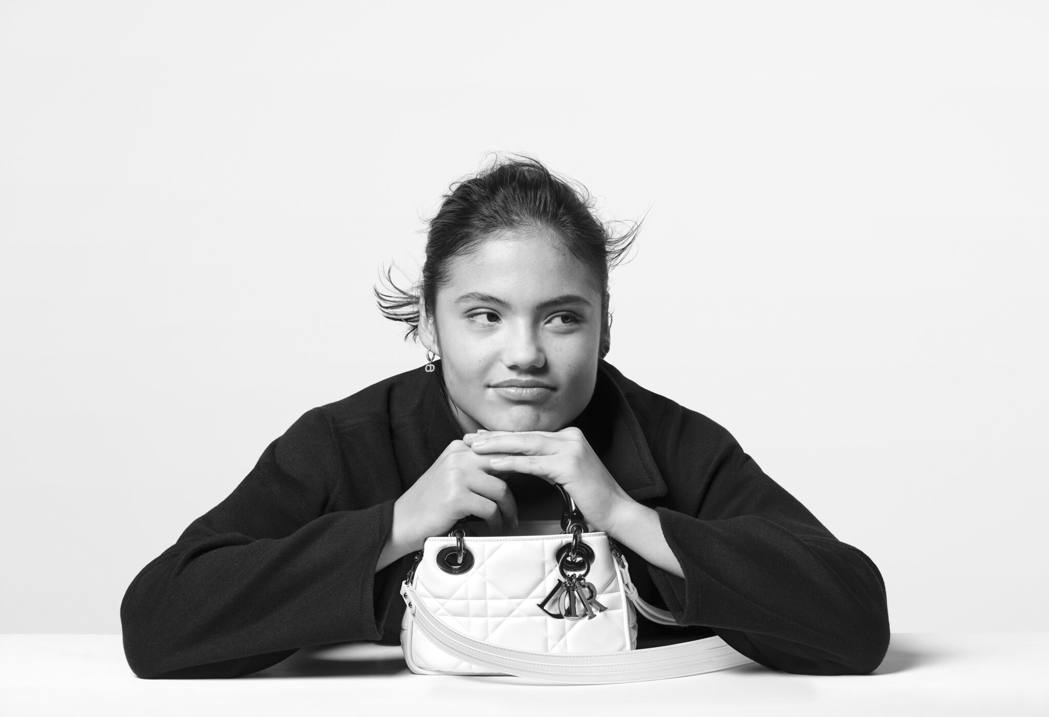 Emma Raducanu, with hands resting on chin, is shown in a studio setting next to a white Lady Dior handbag in this black and white portrait.