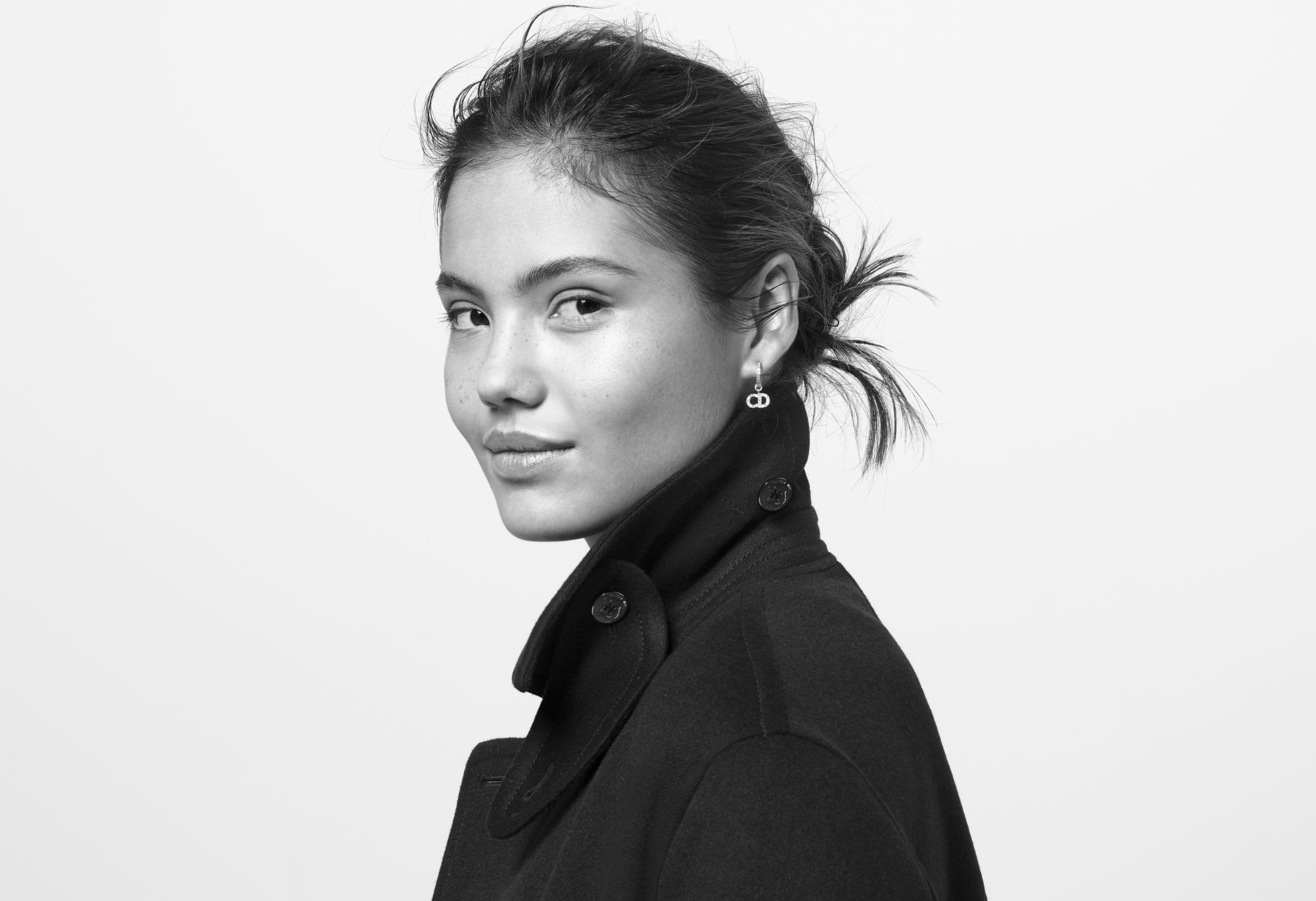 Close-up black and white portrait of a brunette woman wearing a dark coat and earrings, against a bright white backdrop.