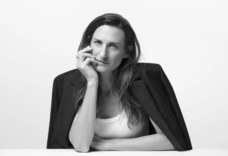 Portrait of adult woman with long hair in white tank top and black blazer, posing indoors against bright white backdrop and studio lighting.