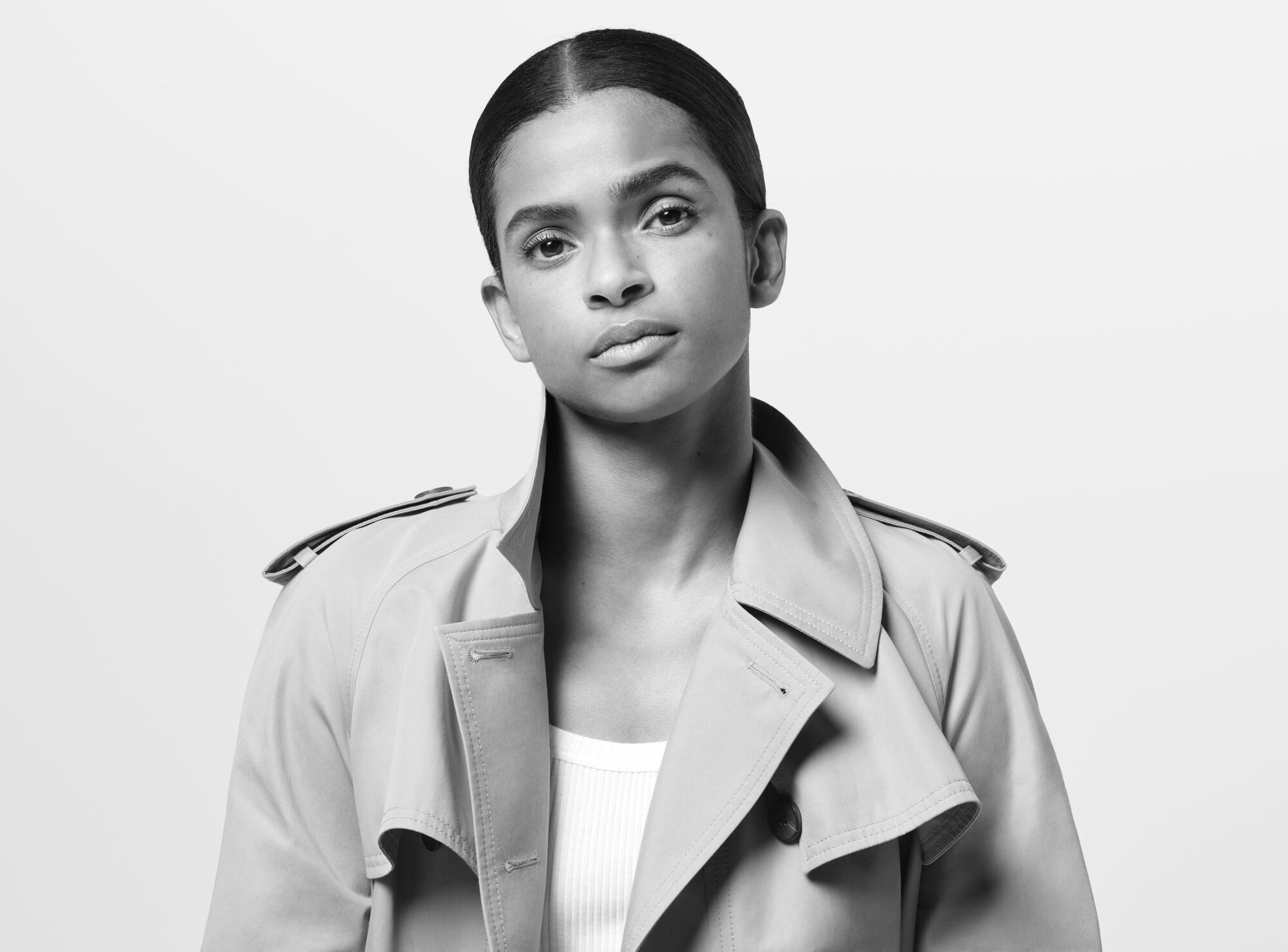 Portrait of an adult woman with slicked back hair in a tan trench coat and white ribbed top against a white studio background.