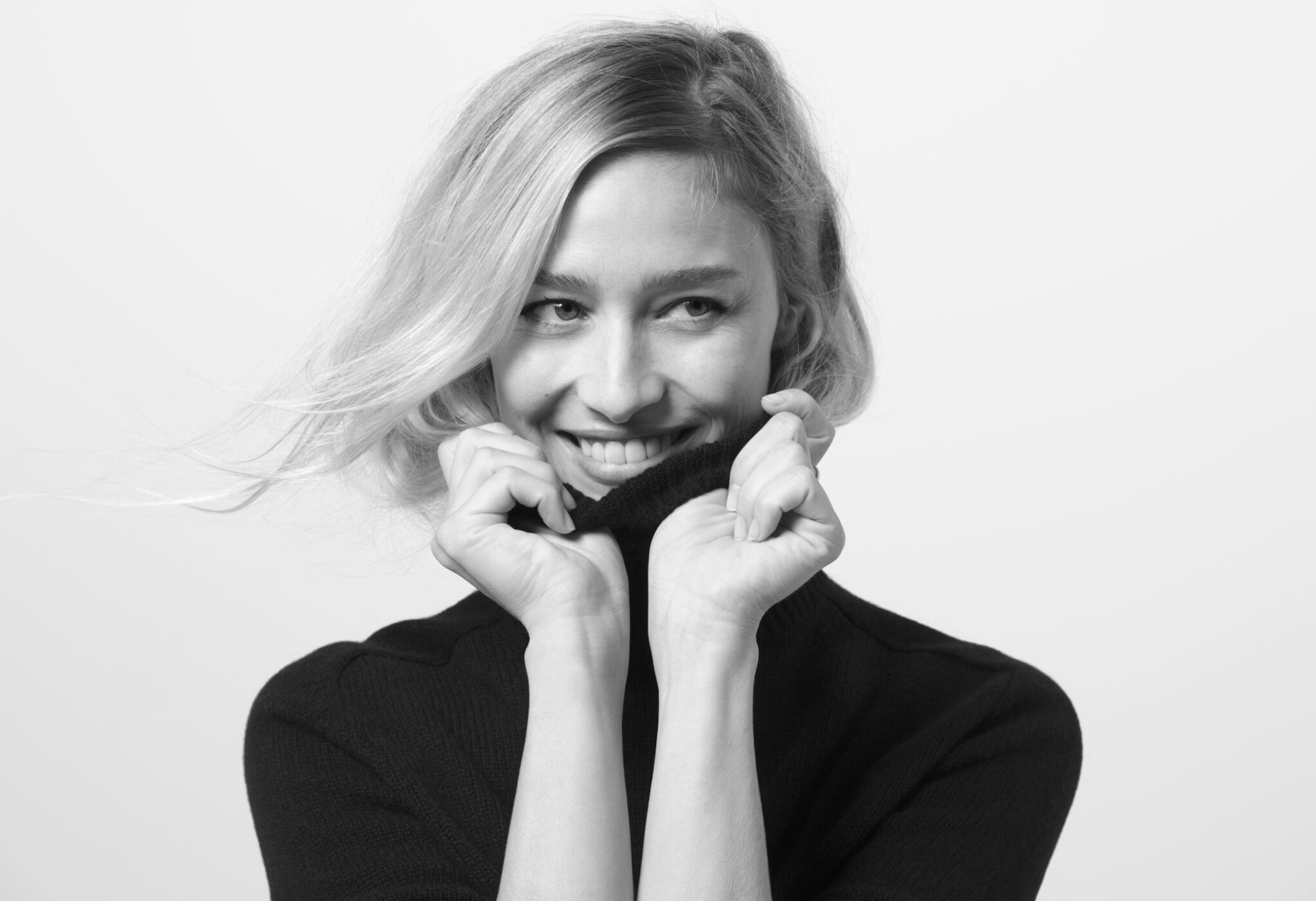 Black and white portrait of a smiling blonde woman in a dark sweater, holding the collar up to her chin in a bright, indoor setting.