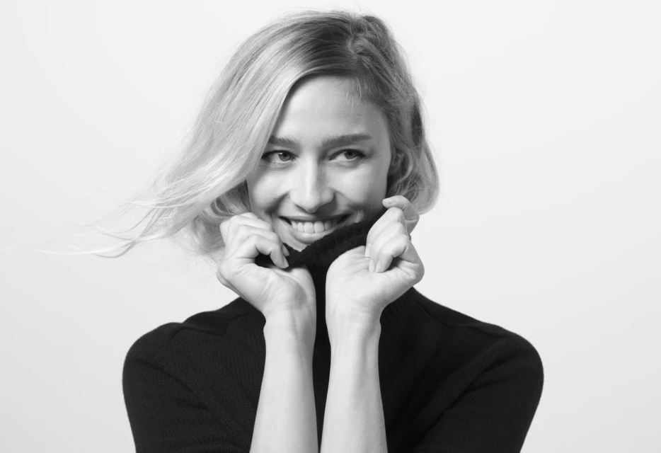 Black and white portrait of a smiling blonde woman in a dark sweater, holding the collar up to her chin in a bright, indoor setting.