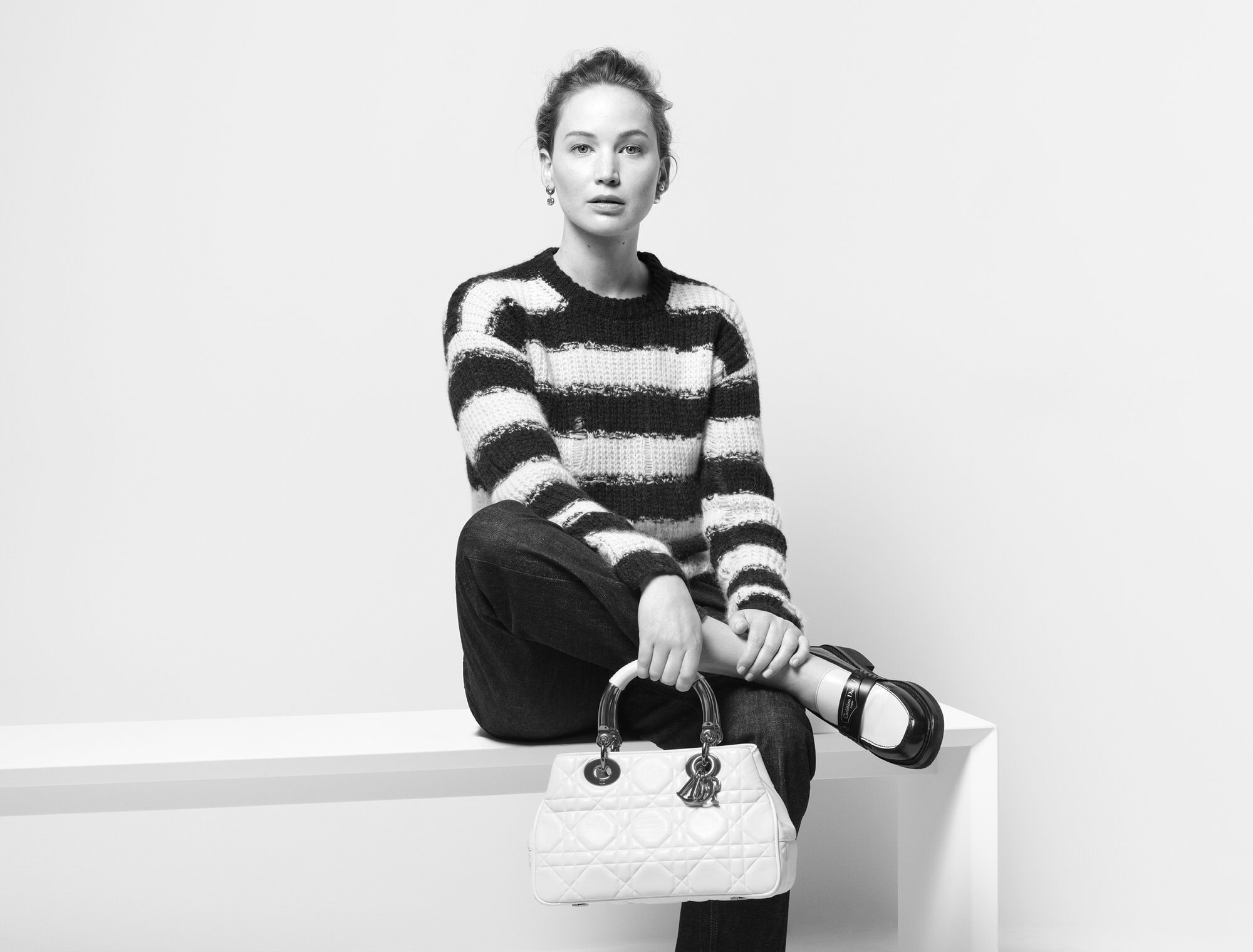 Jennifer Lawrence poses indoors wearing a striped sweater, dark jeans, and black shoes while holding a white Dior handbag.