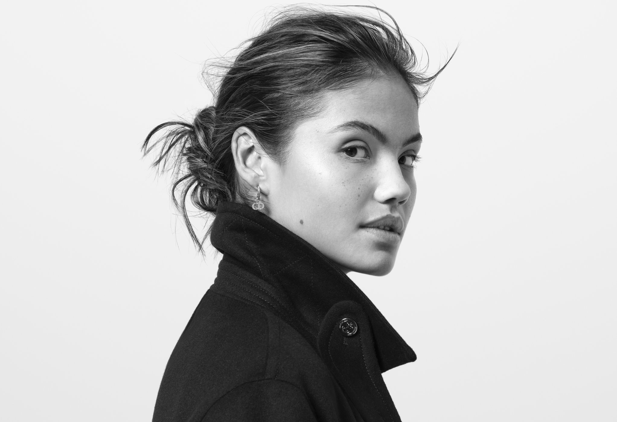 Black-and-white portrait of a woman with her hair in a bun, wearing a dark coat, and looking over her shoulder at eye level.