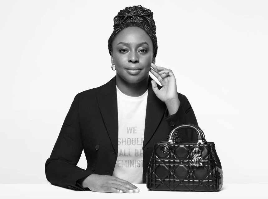 Black and white portrait of a woman with braided hair wearing a blazer, next to a black quilted leather handbag on a white surface.