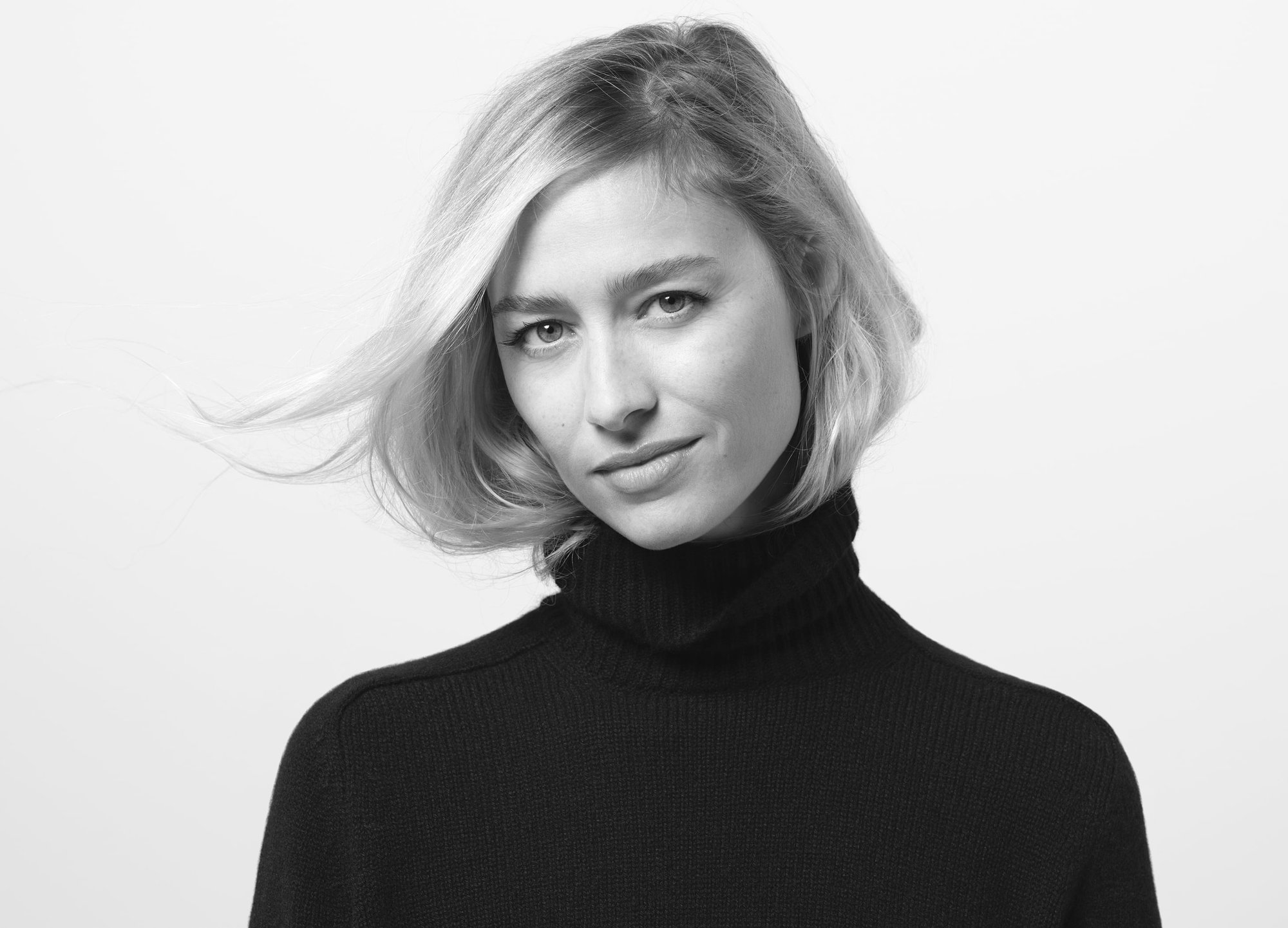 Portrait of a blonde woman with a neutral expression in a black turtleneck sweater against a plain white studio background.