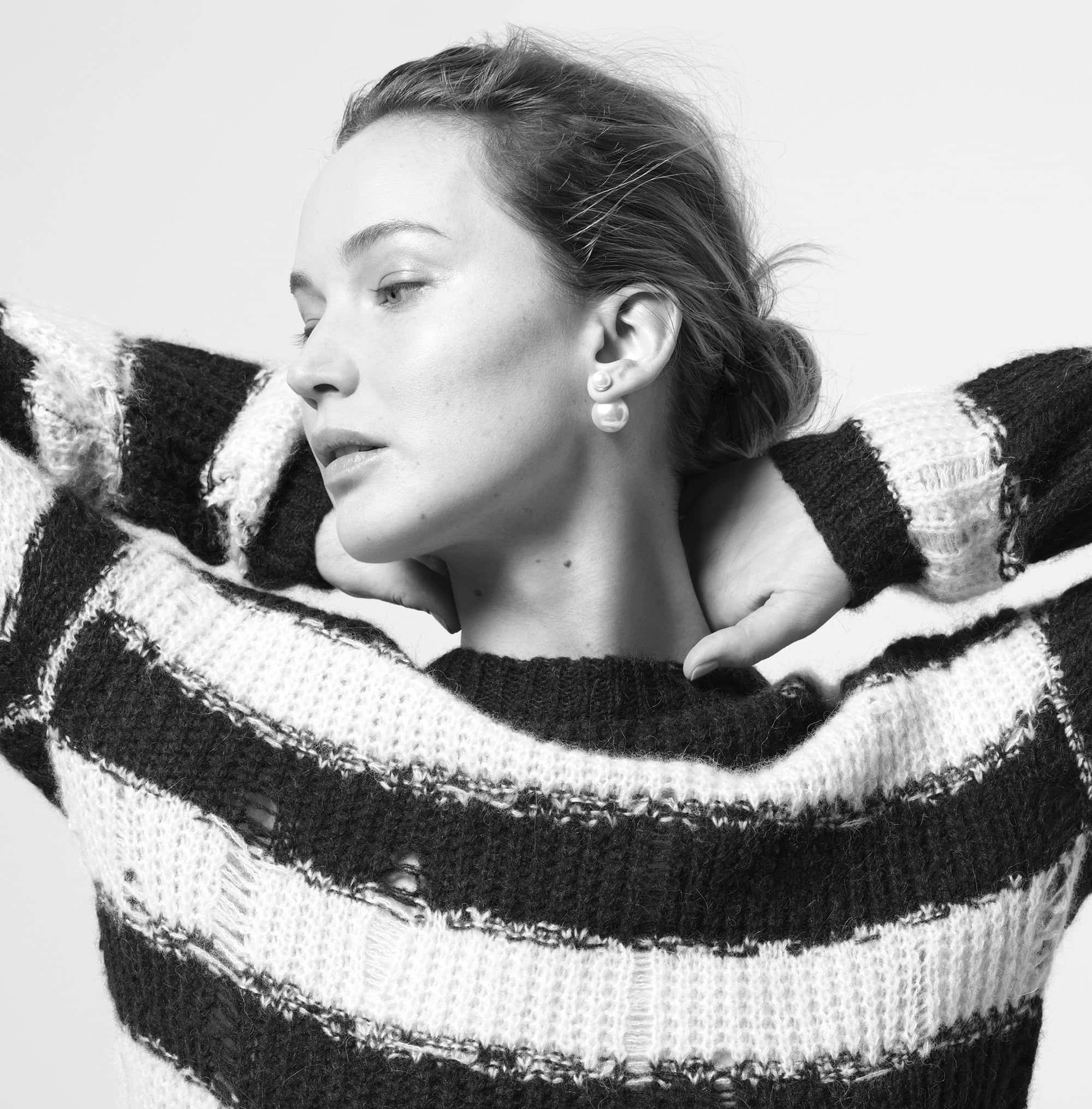 Monochrome portrait of a woman in a striped sweater with a neutral expression, gazing slightly down with hands behind her head.