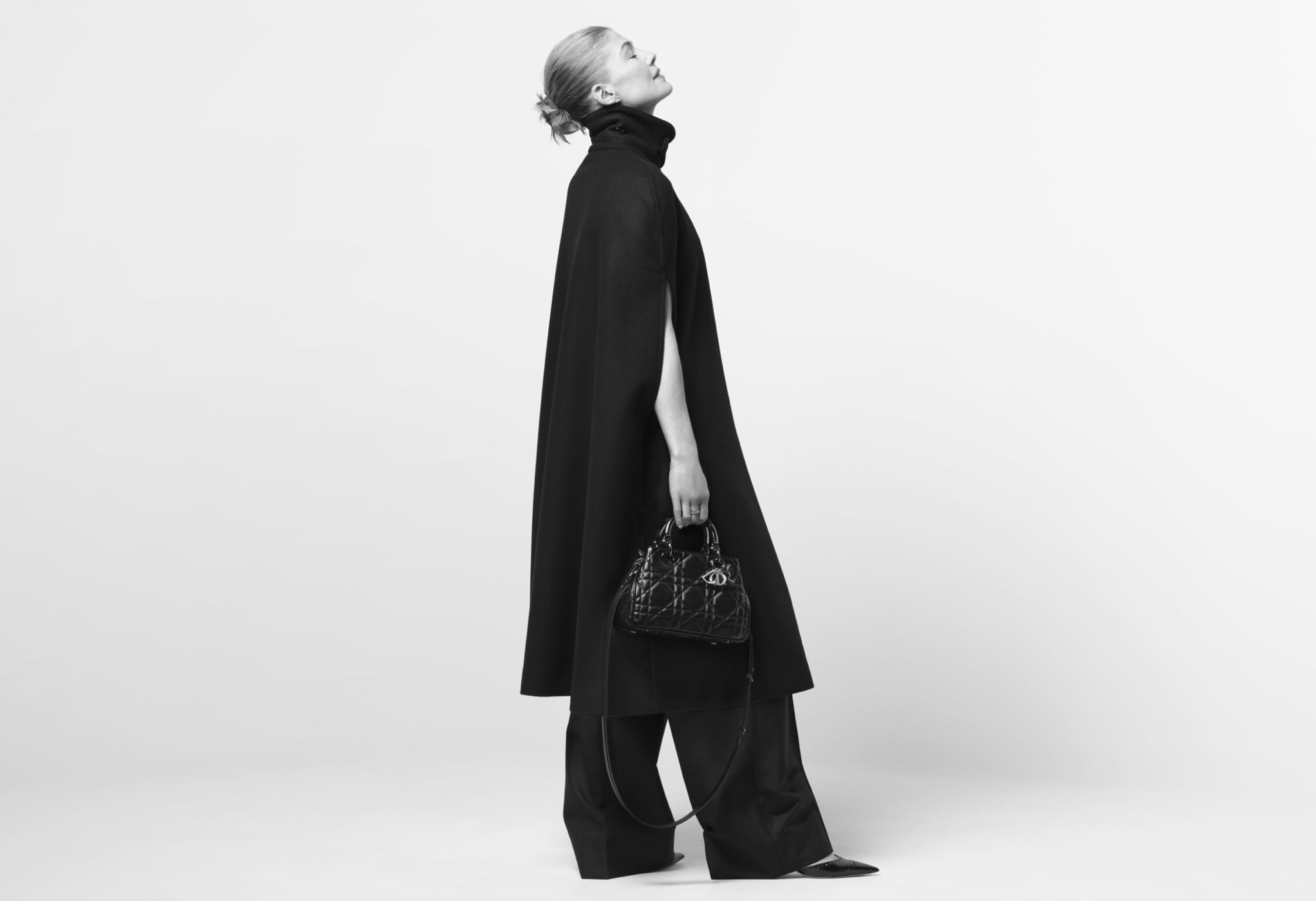 Monochrome fashion shot: Adult woman in black cape and wide-leg pants holds structured handbag in a minimalist photo studio setting.