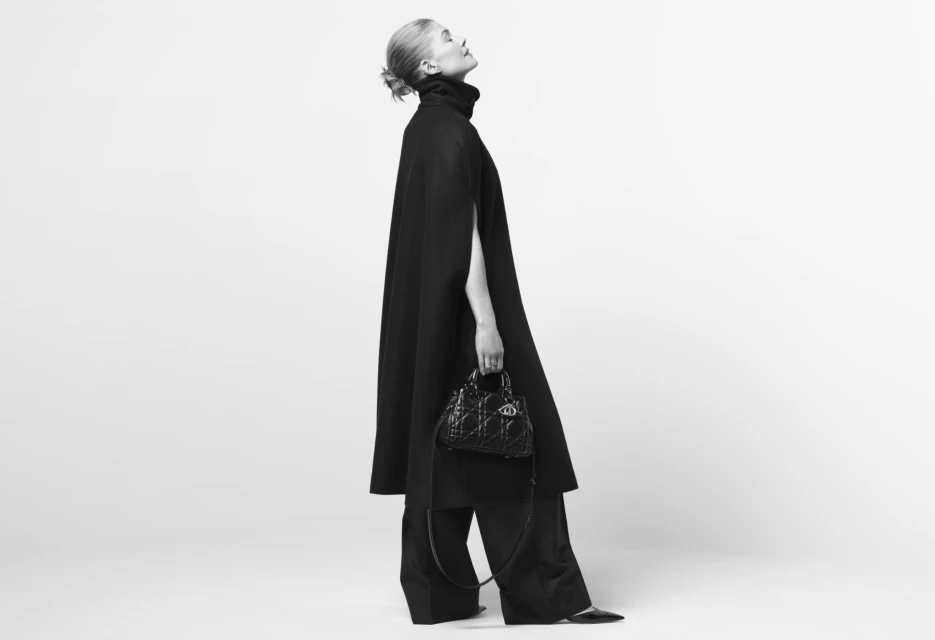 Monochrome fashion shot: Adult woman in black cape and wide-leg pants holds structured handbag in a minimalist photo studio setting.