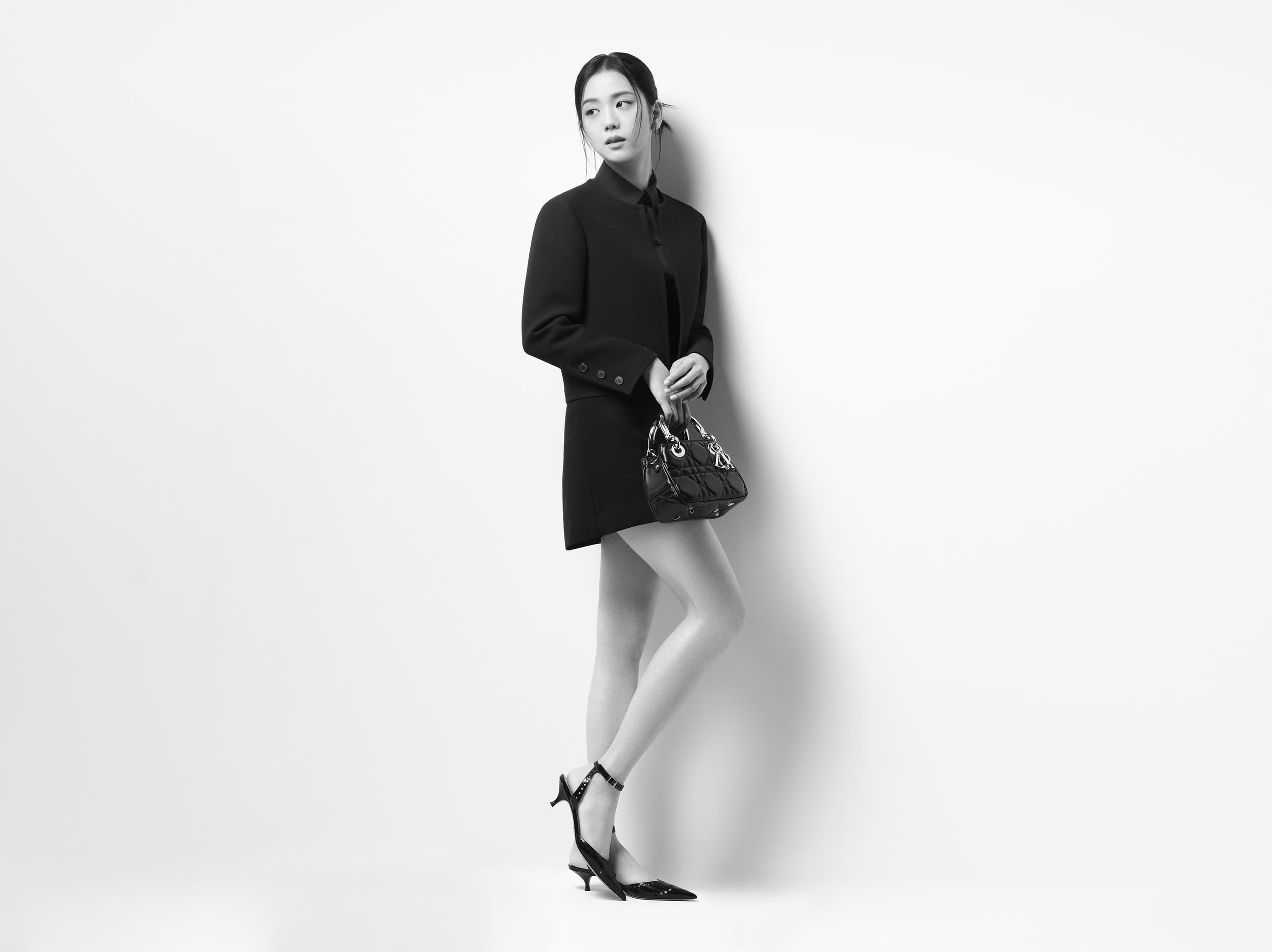 A young Asian woman in a black blazer dress poses in a studio with a black purse and black high heels against a white background.