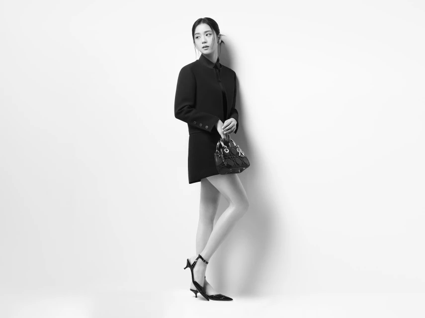 A young Asian woman in a black blazer dress poses in a studio with a black purse and black high heels against a white background.