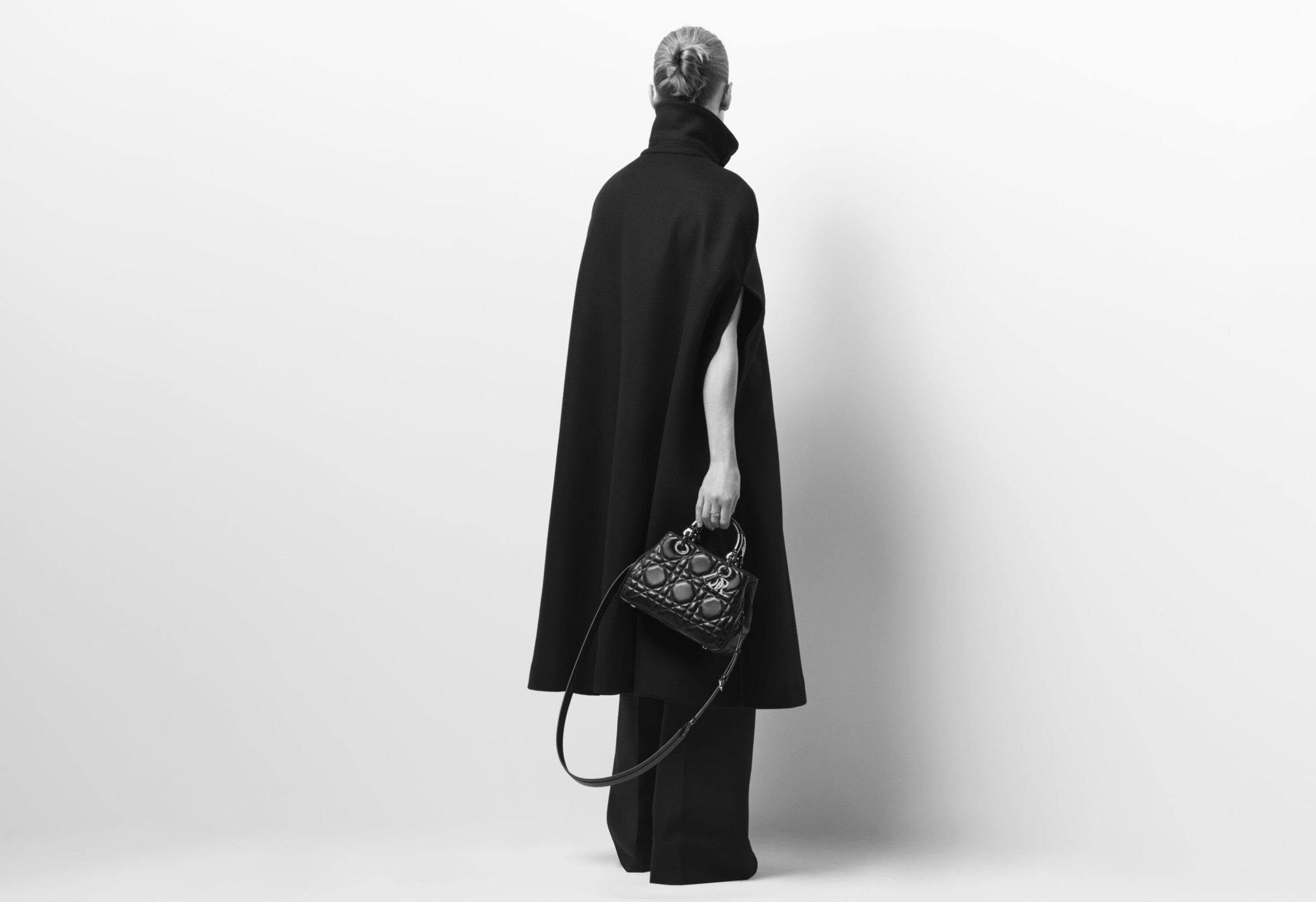 A woman wearing a black cape-like coat and wide-leg pants stands against a white backdrop holding a small, ornate black purse.